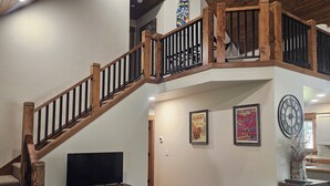 A spacious living area with a wooden staircase, a flat-screen TV on a wooden stand, a black leather chair, and a clock on the wall.
