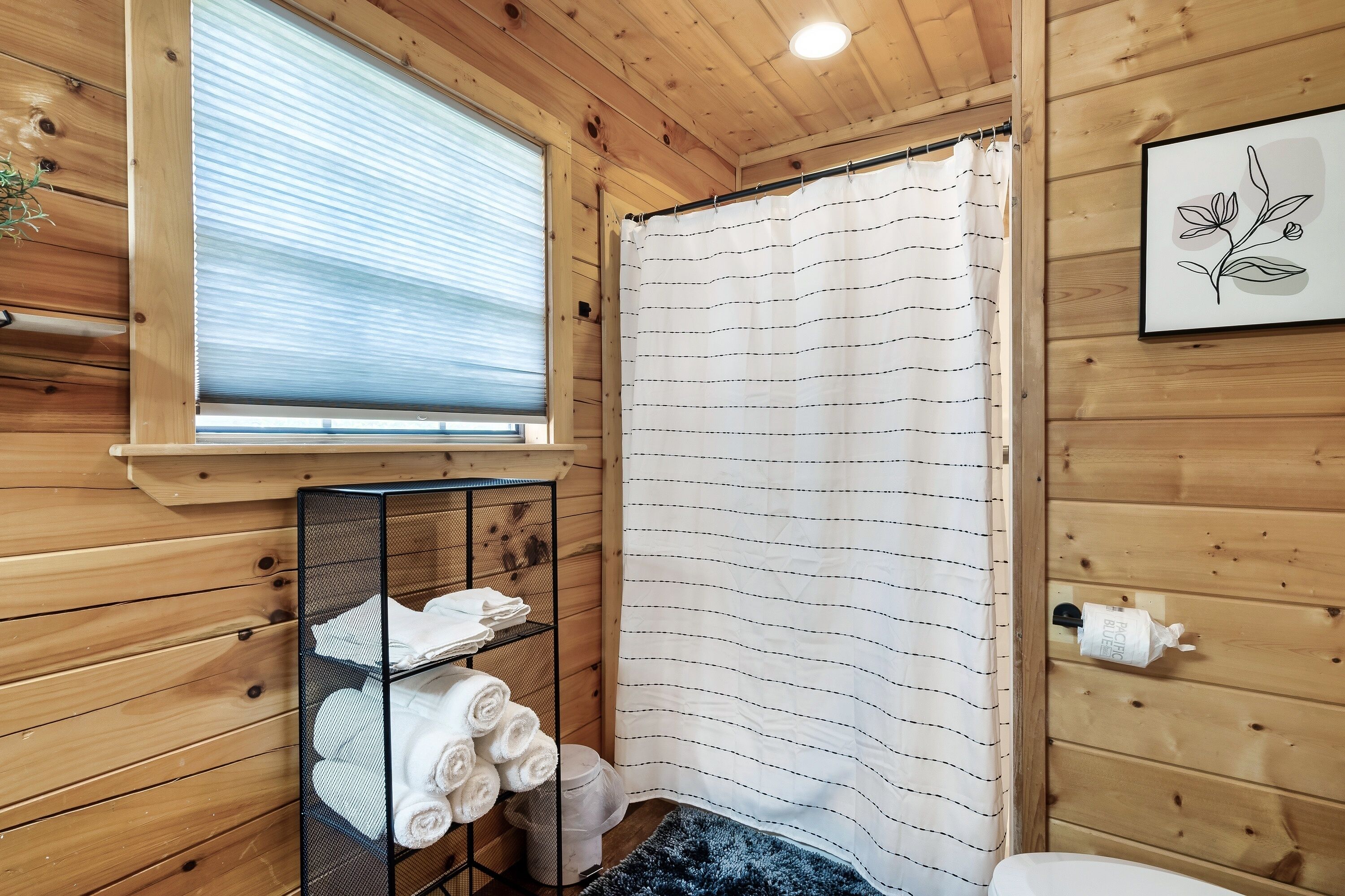 Une salle de bain rustique, avec des murs en bois, comprenant une douche avec rideau, un porte-serviettes avec des serviettes, une fenêtre et un tableau encadré accroché au mur.