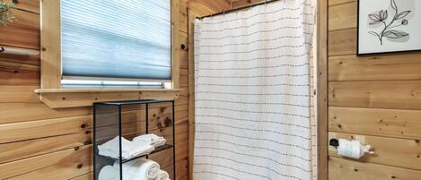 Une salle de bain rustique, avec des murs en bois, comprenant une douche avec rideau, un porte-serviettes avec des serviettes, une fenêtre et un tableau encadré accroché au mur.