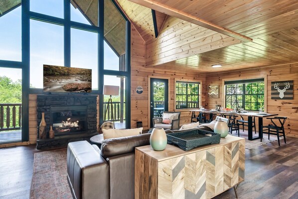 A spacious cabin with a fireplace, leather seating, and large windows offering views of the outdoors. - Renovated Cabin. Hot tub, Arcade, Pool Table, Shuffle Board, Dollywood, Parking  (Sevierville)