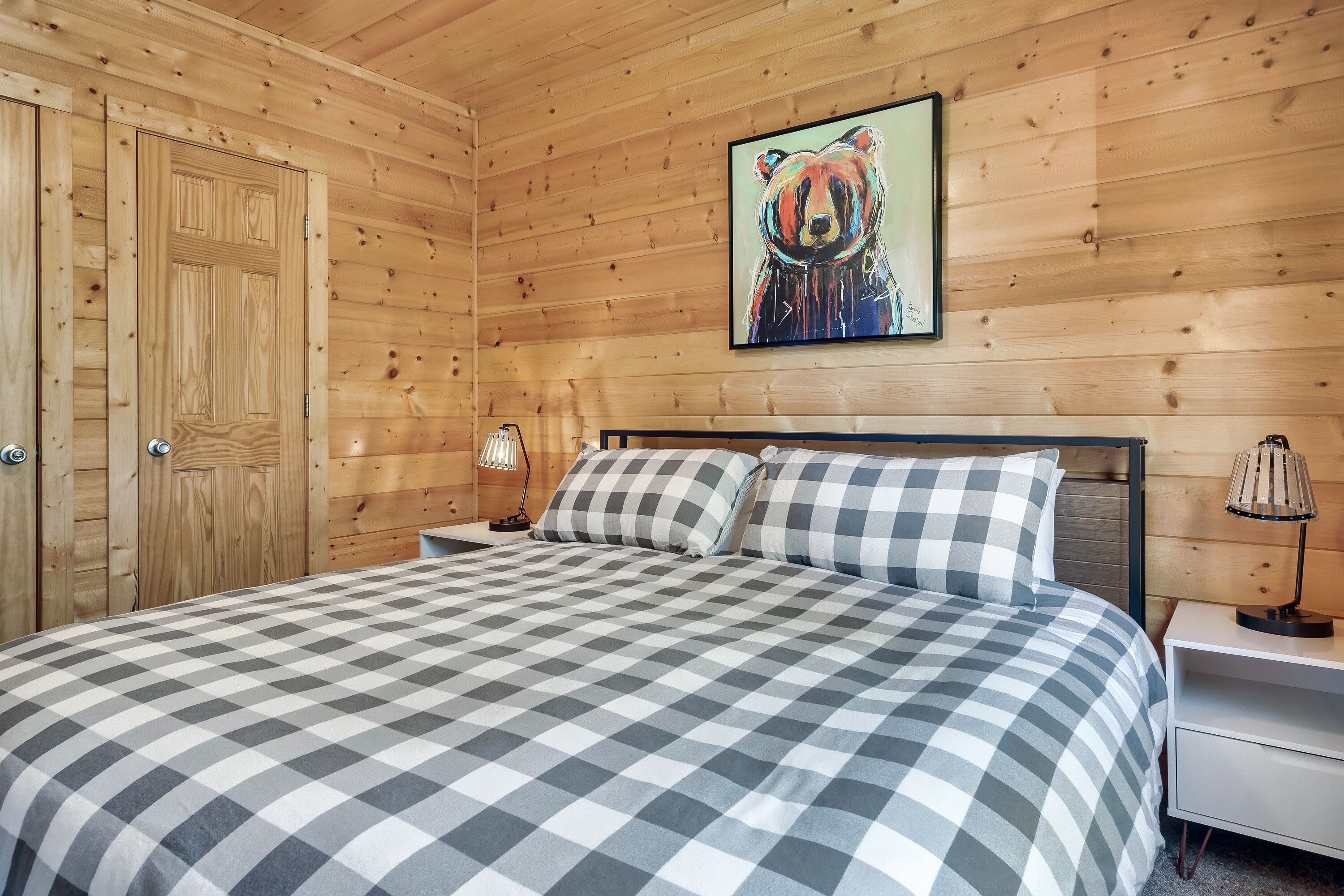 A wooden-paneled room with a bed, checkered bedding, a painting of a bear, and a nightstand with a lamp.