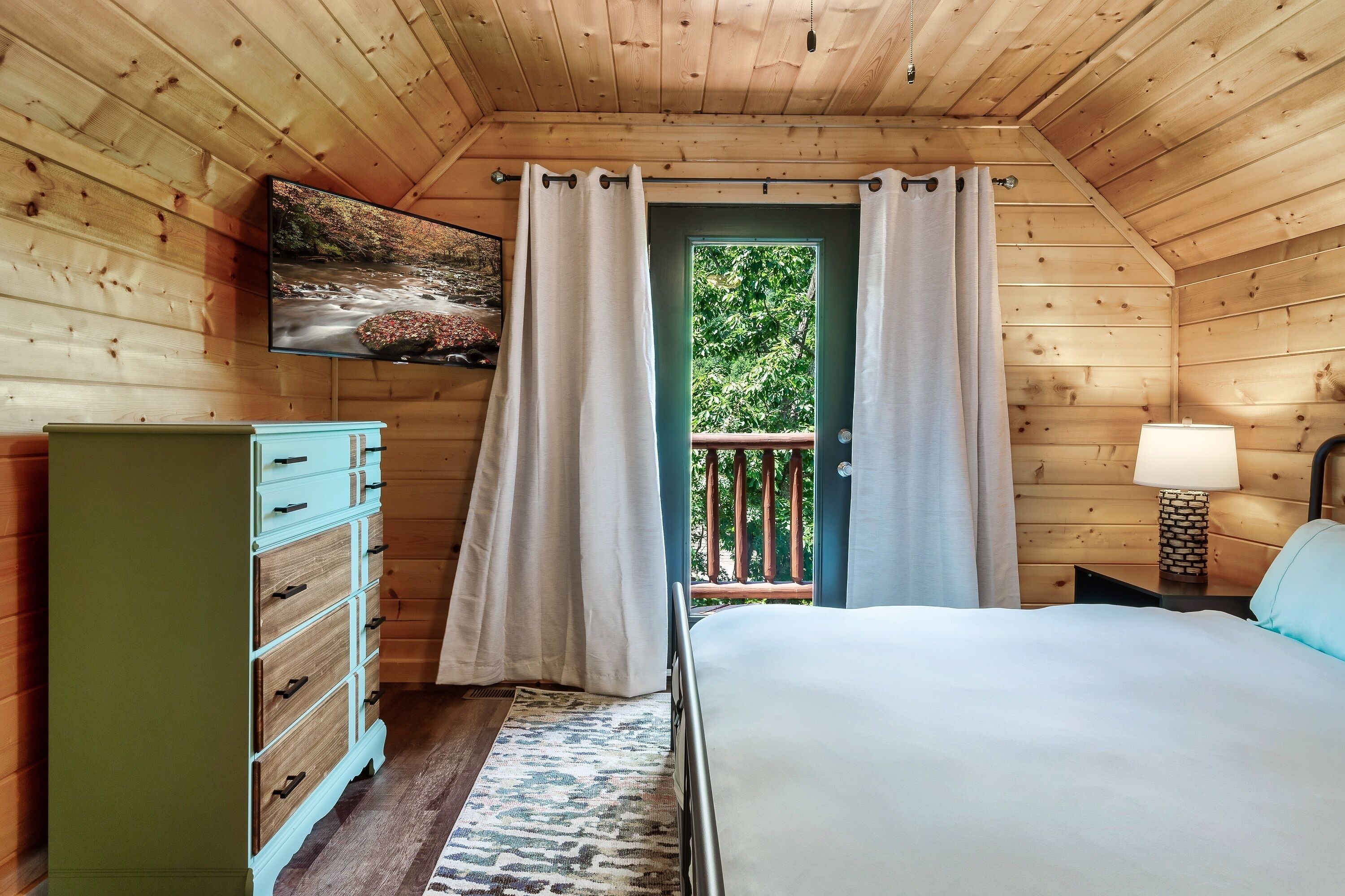 A wooden cabin bedroom with a bed, dresser, television, and a balcony with curtains.