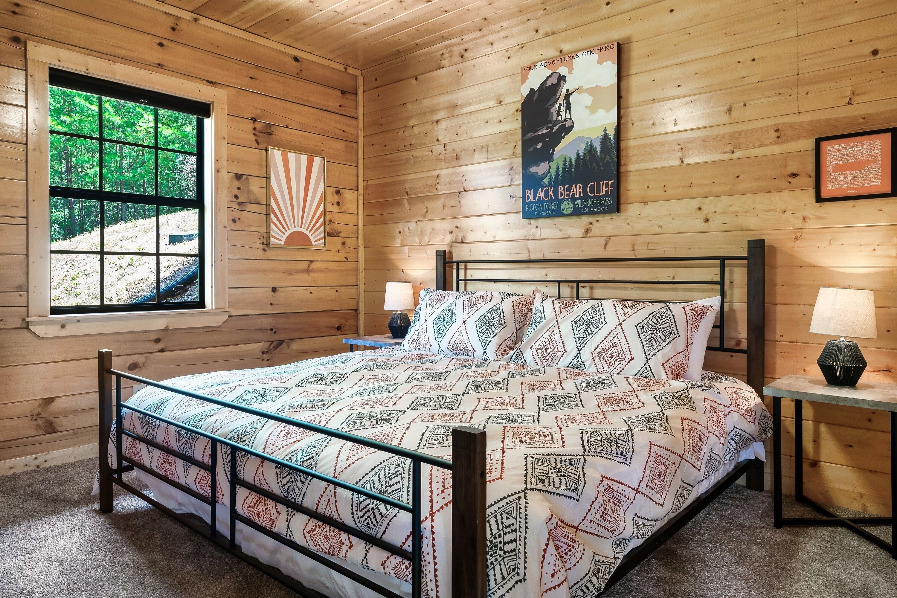 A wooden cabin room with a bed, two bedside lamps, a poster on the wall, and a window with a view of trees.