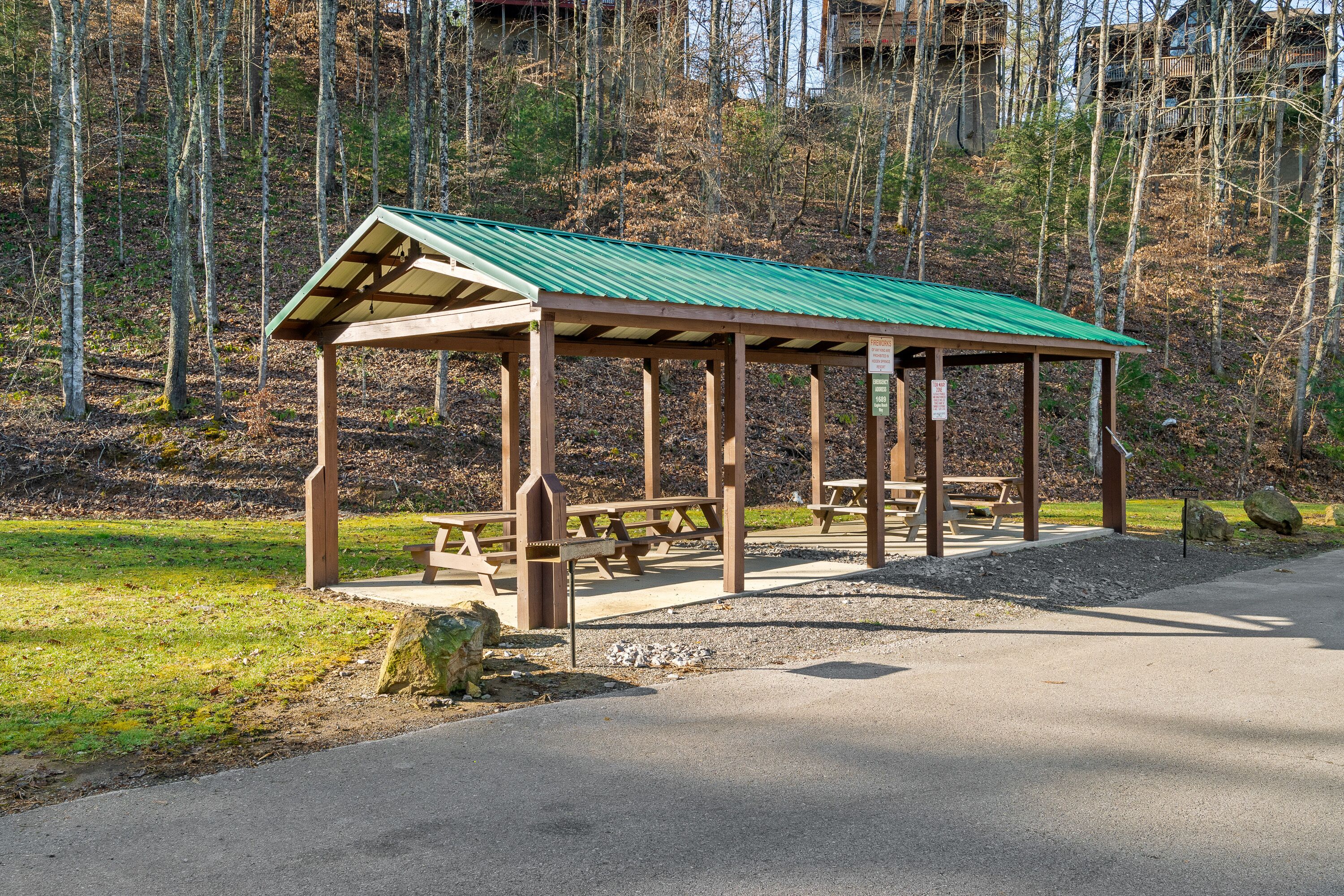 A wooden pavilion with a green roof, picnic tables, and benches in a forested area.