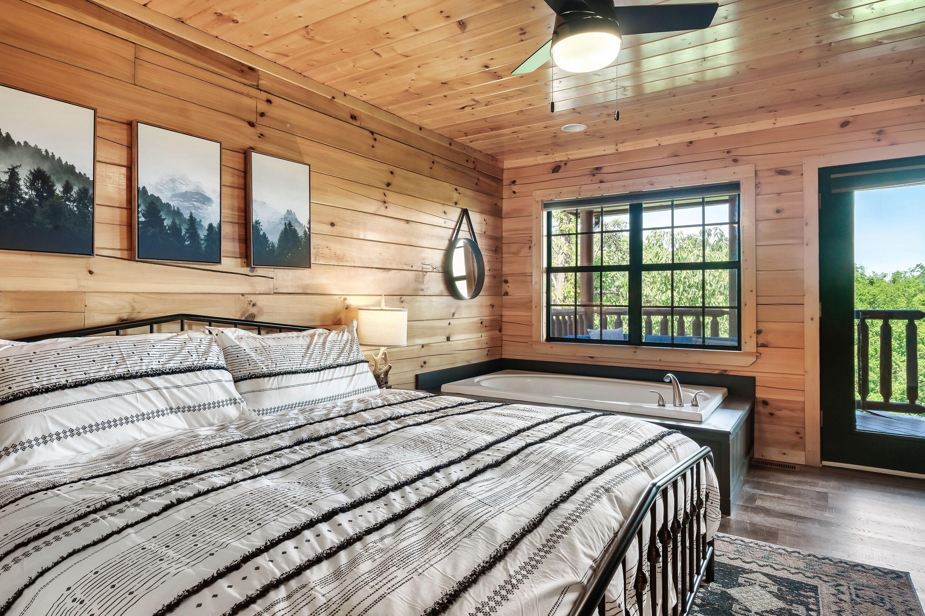 A wooden cabin bedroom with a large bed, a window with a view of greenery, and a small bathroom with a sink and bathtub.