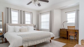 A bedroom with a bed, a desk, a chair, a ceiling fan, and large windows with curtains.