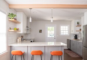 Una cocina moderna con armarios blancos, una isla central con taburetes de bar, iluminación colgante y vistas al exterior.