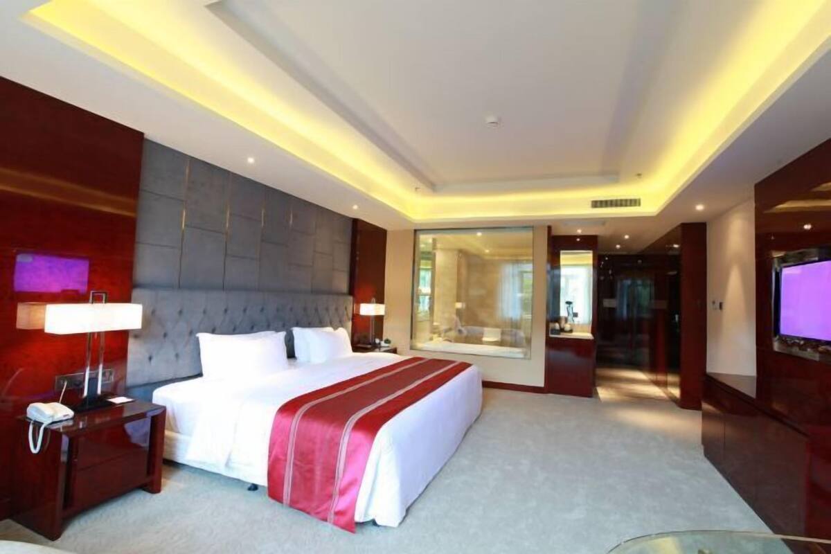 A modern hotel room with a large bed, bedside tables, a TV, and a glass door leading to a bathroom.
