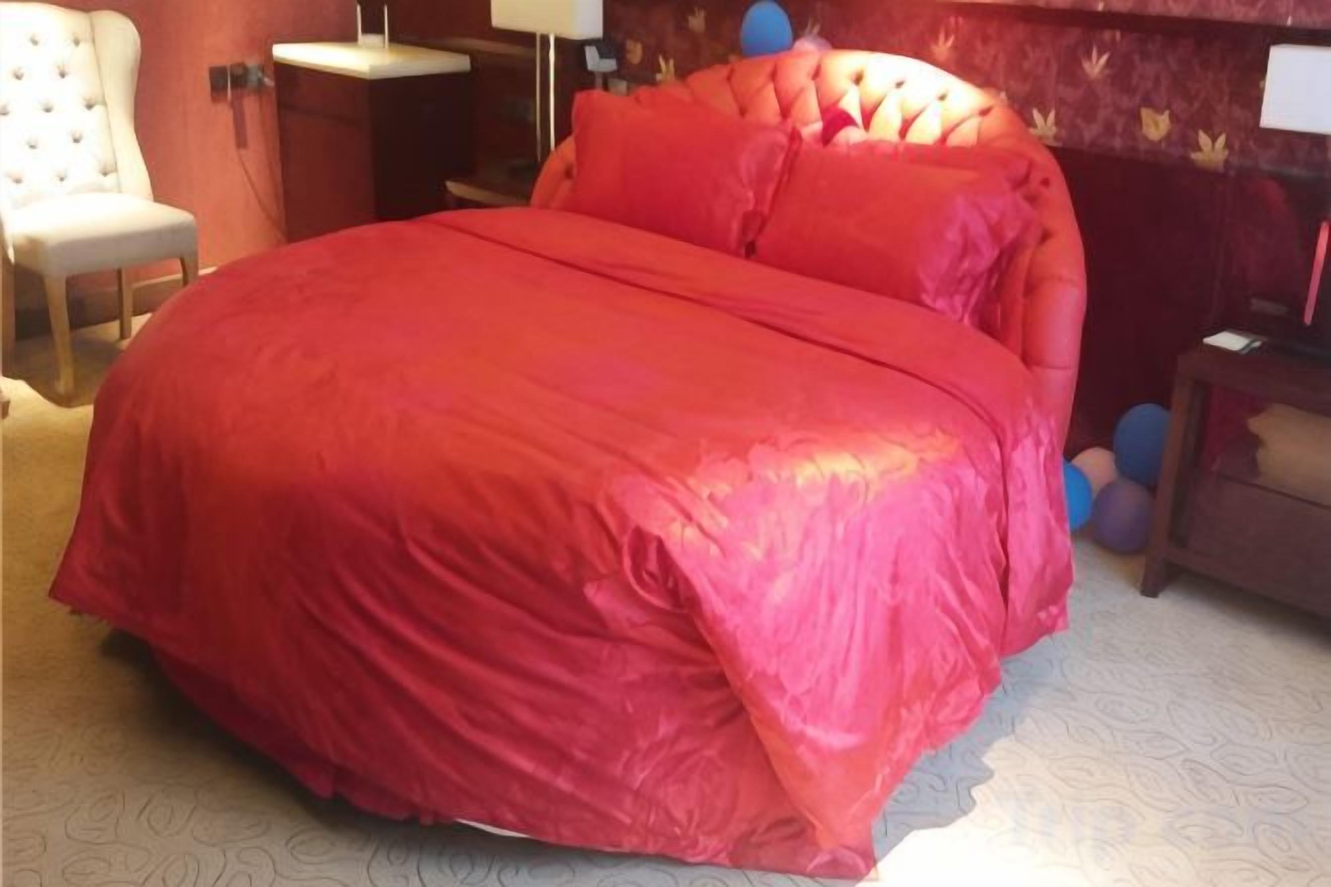 Una cama con colcha roja, una silla, una mesita de noche y una lámpara.