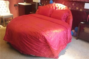 Una cama con colcha roja, una silla, una mesita de noche y una lámpara.