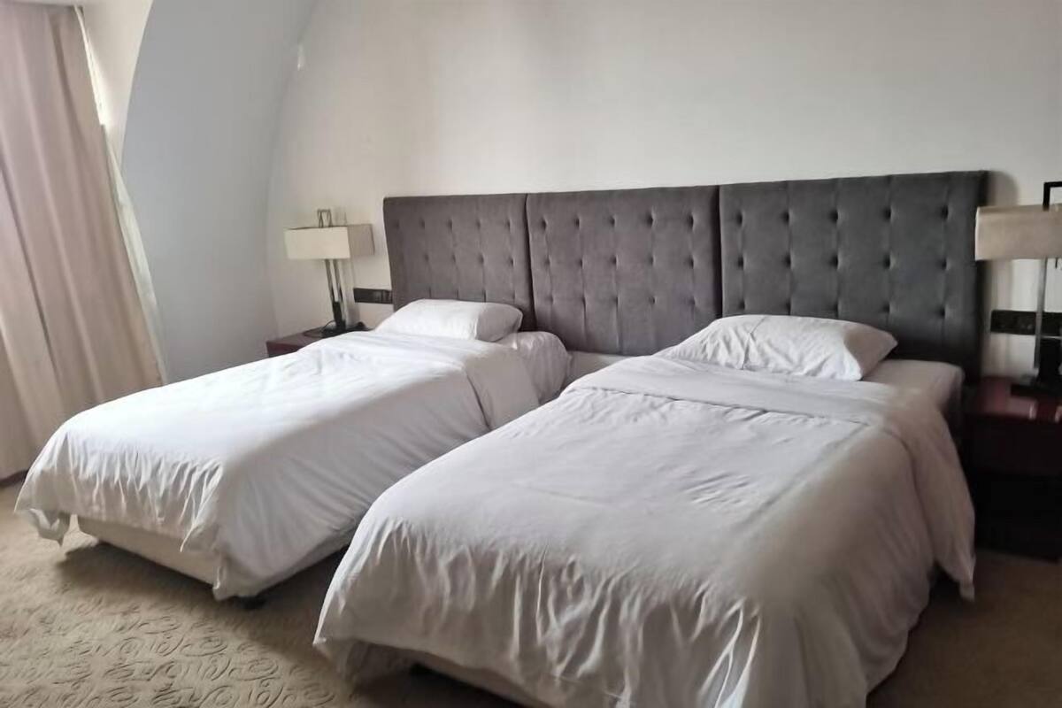 A hotel room with two beds, a grey upholstered headboard, white bedding, and a lamp on each side.
