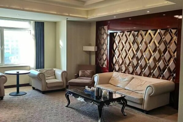 Quarto de hotel com sofá bege, duas poltronas, uma mesinha redonda e um painel decorativo na parede.