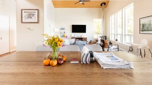 Ein modernes Wohnzimmer mit Esstisch aus Holz, einer Couch und einem Fernseher. Fenster sorgen für natürliches Licht und ein Deckenventilator ist vorhanden.