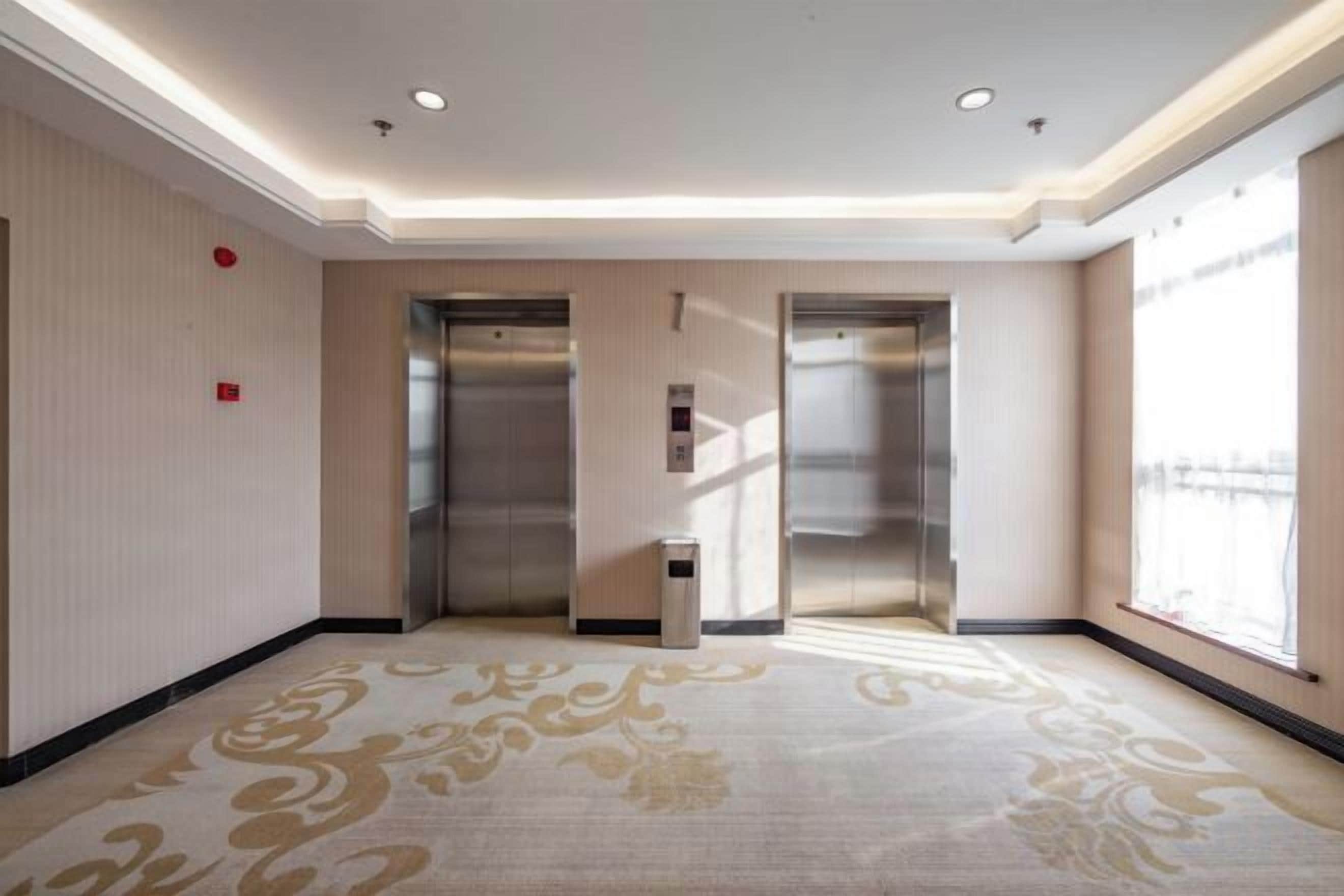 A hotel corridor with two elevators, a patterned carpet, and a large window on the right.