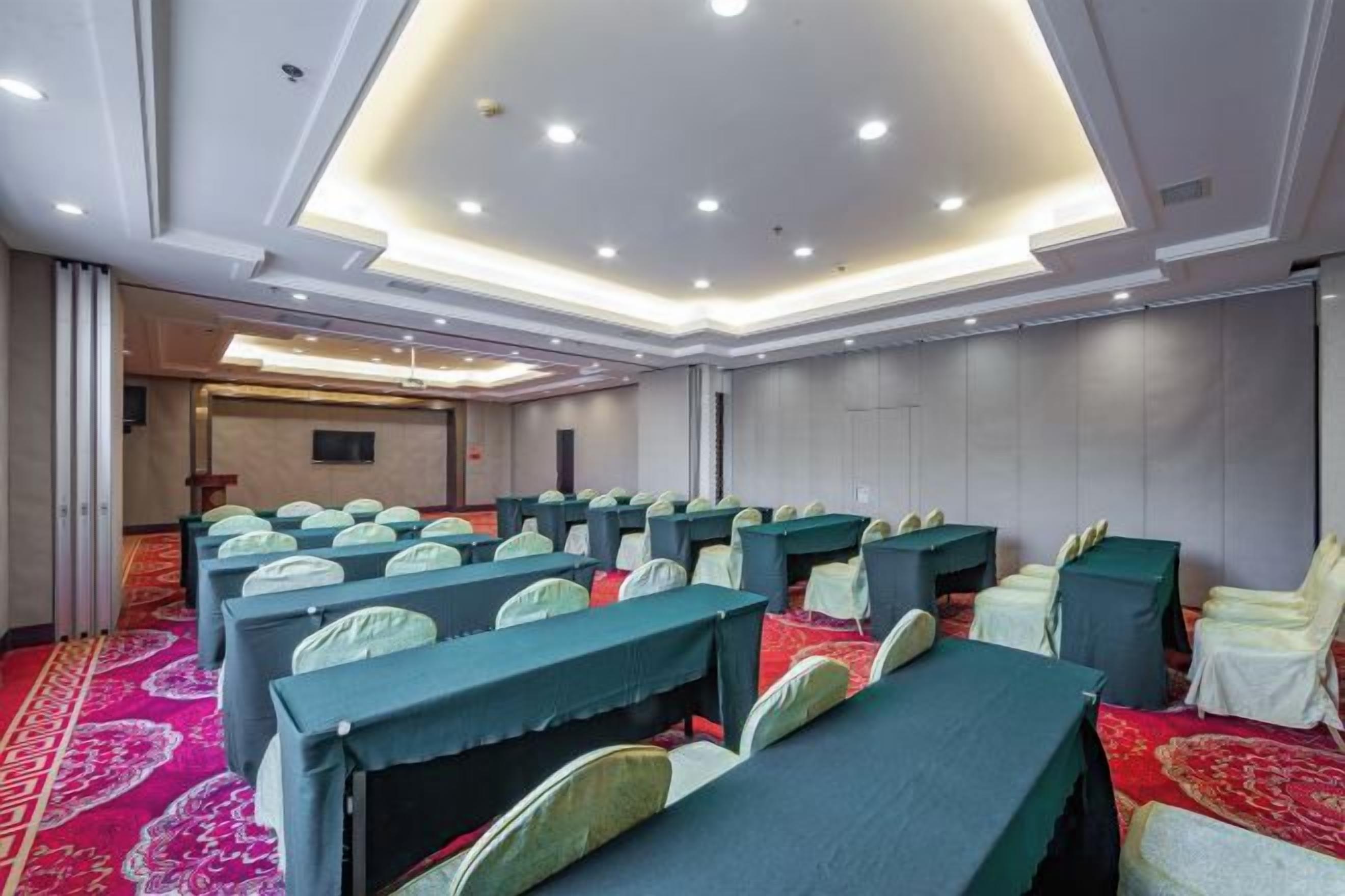 Un salón de conferencias con filas de mesas y sillas, piso alfombrado e iluminación empotrada en el techo.