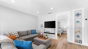 A modern living room with a grey sofa, ottoman, and a flat-screen TV mounted on the wall. - Walk to Beach. Beautifully Renovated 3BR, 2BA Home w/Outdoor Shower & Grille (Nokomis)