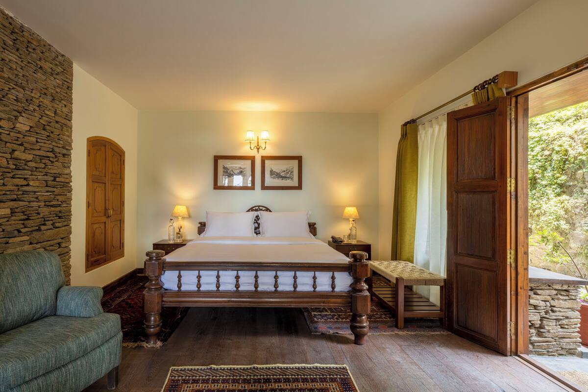 A bedroom with a large bed, wooden headboard, two bedside lamps, a stone wall, a wooden door, and a bench.