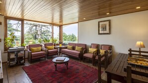 Una sala de estar acogedora con muebles de madera, un tapete rojo y grandes ventanales que dan a zonas verdes.