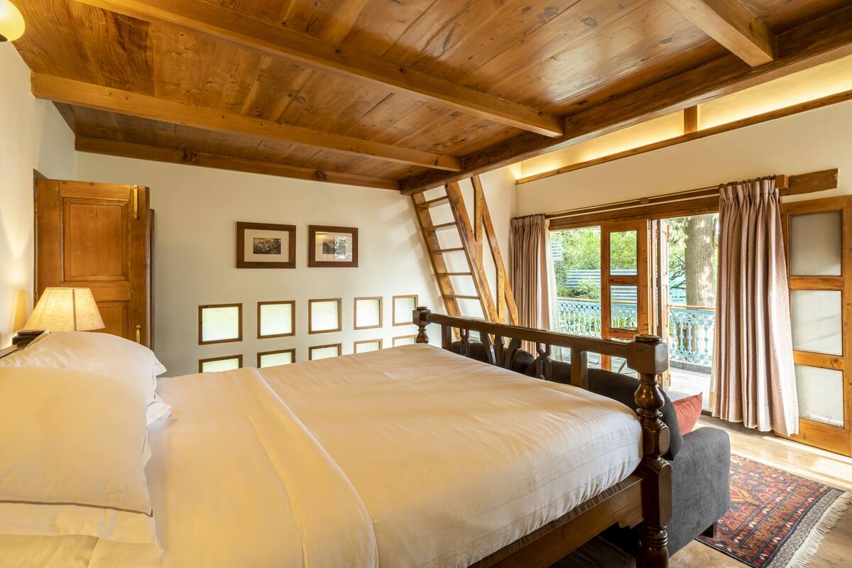 A bedroom with a wooden ceiling, a bed with white linens, a wooden ladder, and a view of greenery outside.