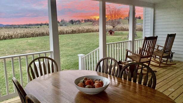 A covered patio with a wooden table and chairs, a bowl of fruit, and a view of a field and sunset. - 1875 FARMHOUSE renovated with a spectacular view! SUMMER SPECIALS! (Luray)