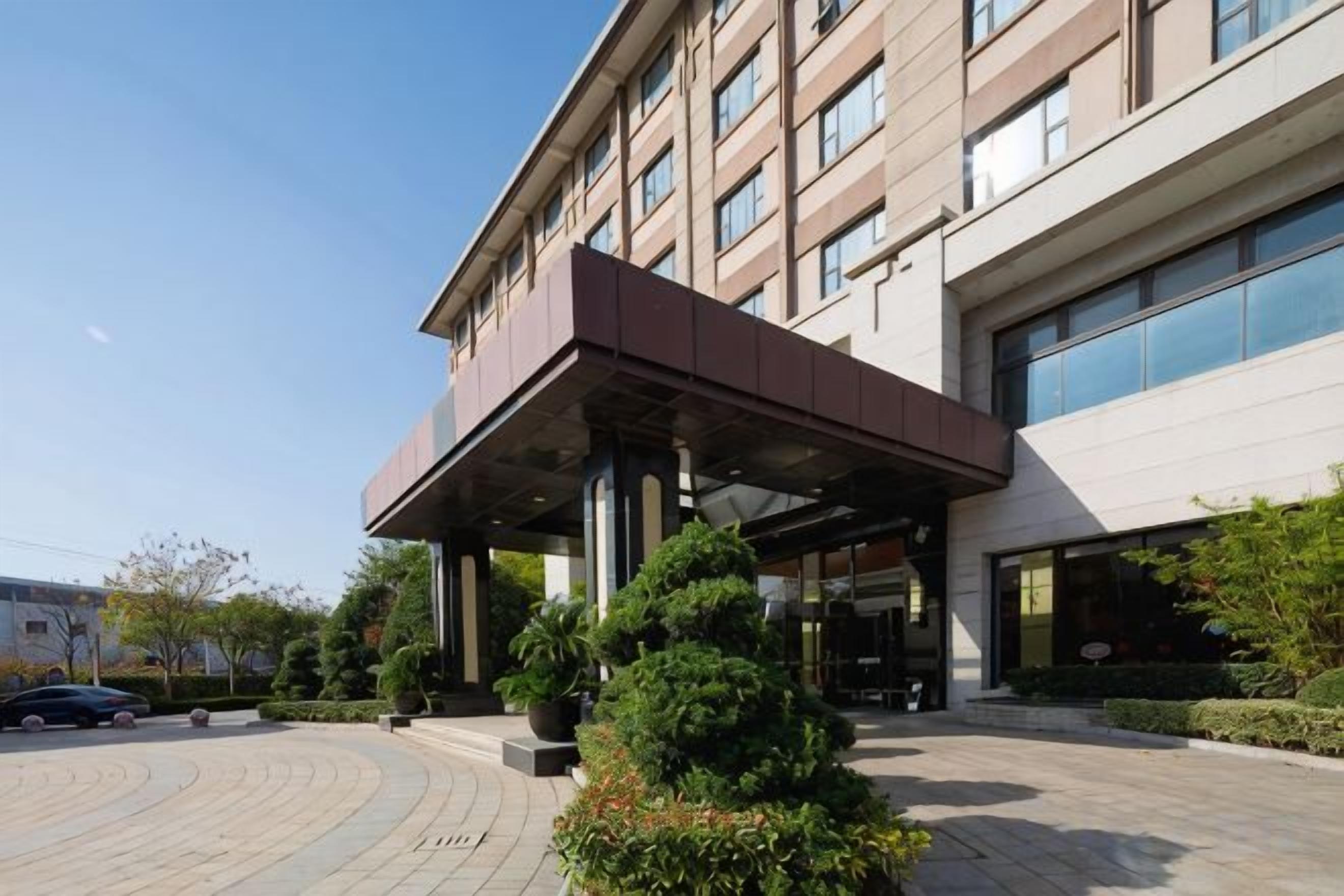 A modern hotel entrance with a covered walkway, large windows, and a landscaped area with shrubs.