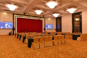 Un salón de conferencias con filas de sillas, un escenario con cortina roja y una pantalla grande.