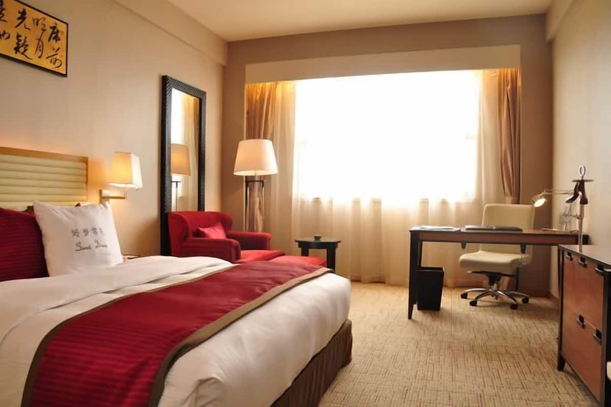 A hotel room with a bed, a desk, a chair, a lamp, and a window with curtains.