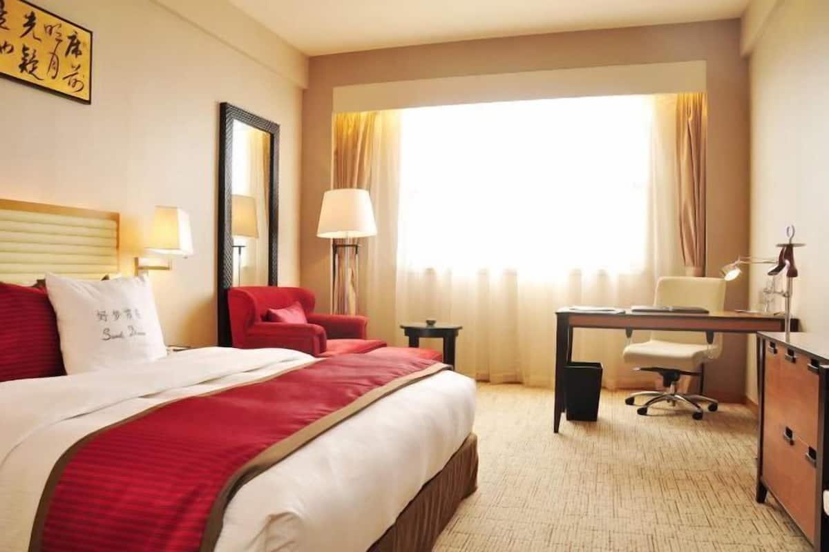 A hotel room with a bed, a desk, a chair, a lamp, and a window with curtains.