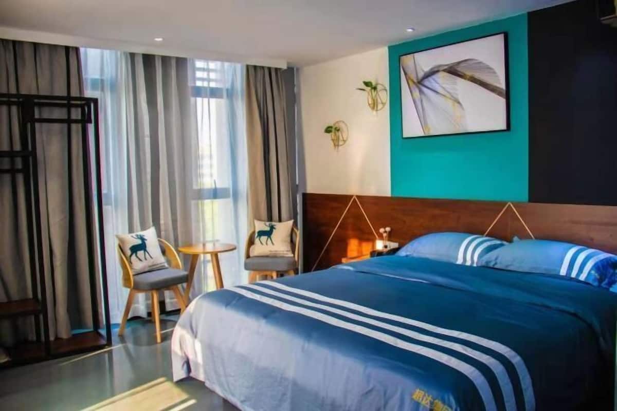 Habitación de hotel con una cama grande, una mesita, dos sillas, un ventanal con cortinas y una obra de arte colgada en la pared.