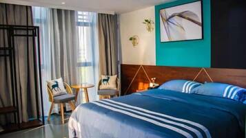 Ein Hotelzimmer mit einem großen Bett, einem kleinen Tisch, zwei Stühlen, einem Fenster mit Vorhängen und einem an der Wand befestigten Kunstwerk.