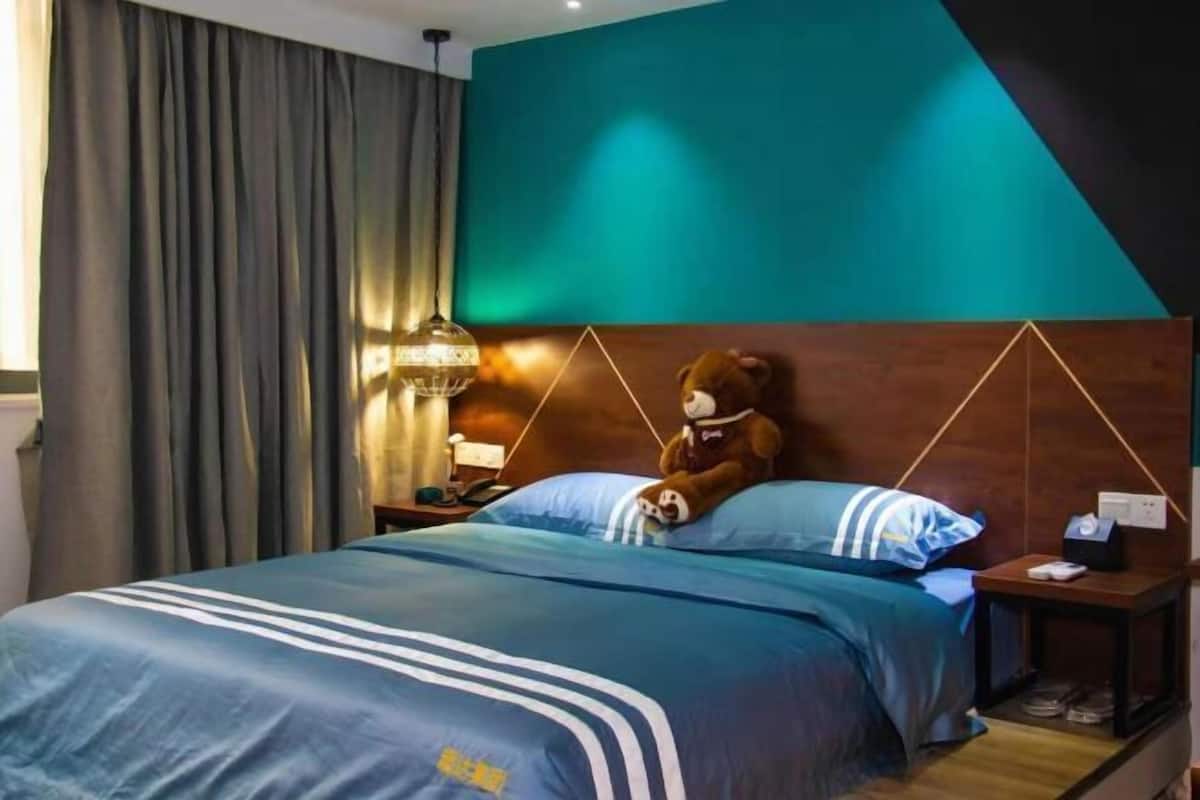 Una habitación de hotel con una cama grande, un cabecero de diseño geométrico, una mesita de noche con una lámpara y un osito de peluche sobre la cama.