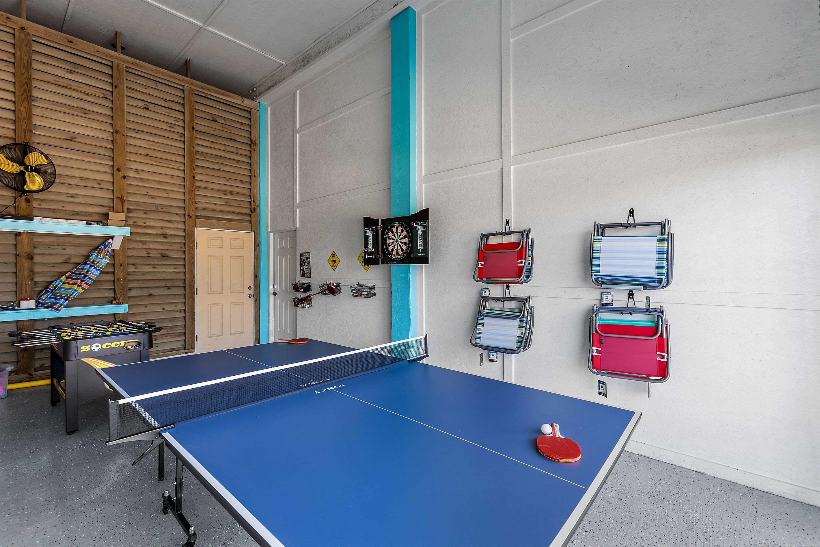 A ping pong table with a blue table and net, a foosball table, and a dartboard in a room with wooden paneling and a white door.