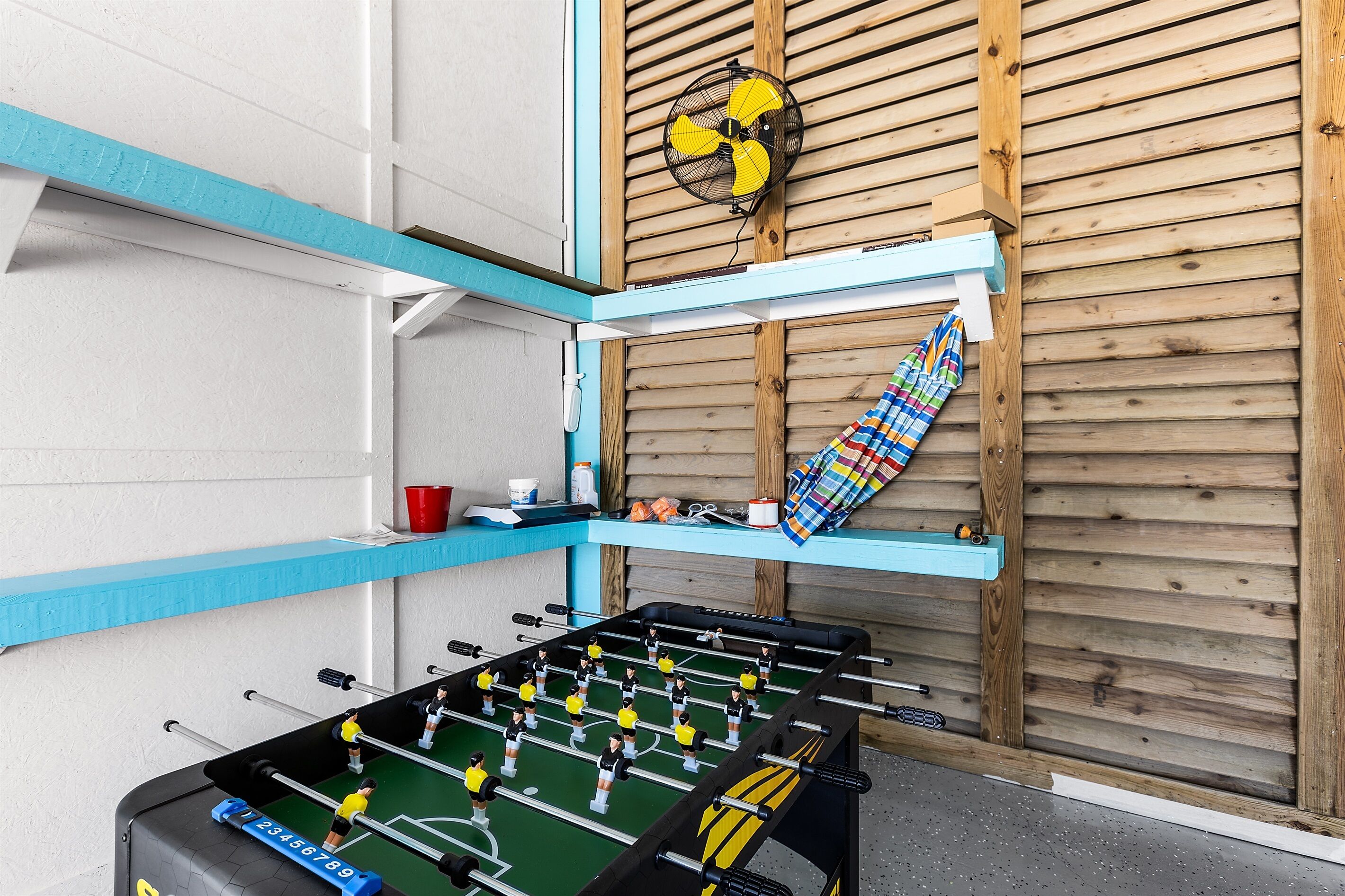 A foosball table in a room with wooden panel walls, a fan, and a shelf with various items.