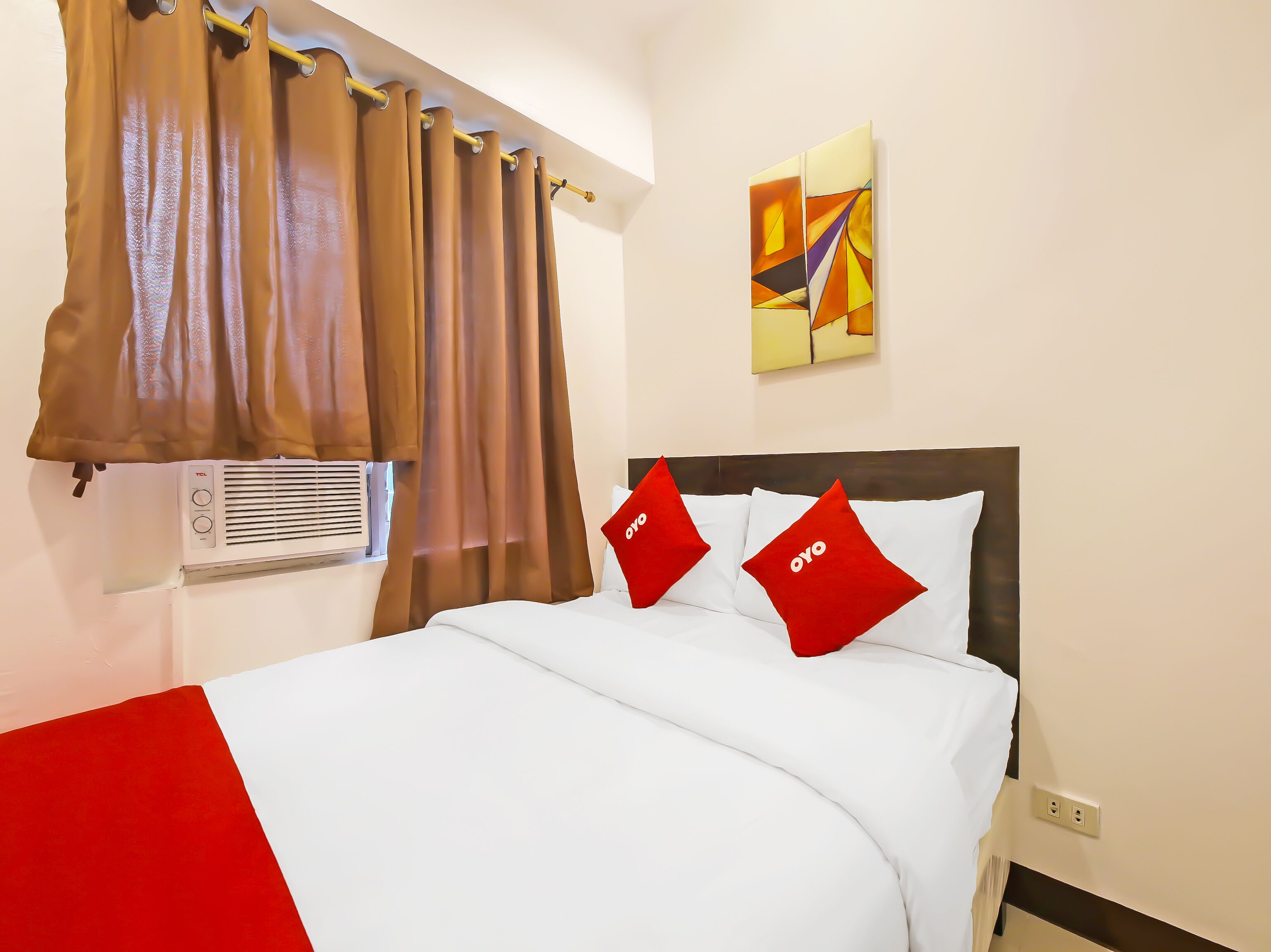 Una habitación de hotel con cama, ropa de cama roja y blanca, ventana con cortinas, equipo de aire acondicionado y un cuadro en la pared.