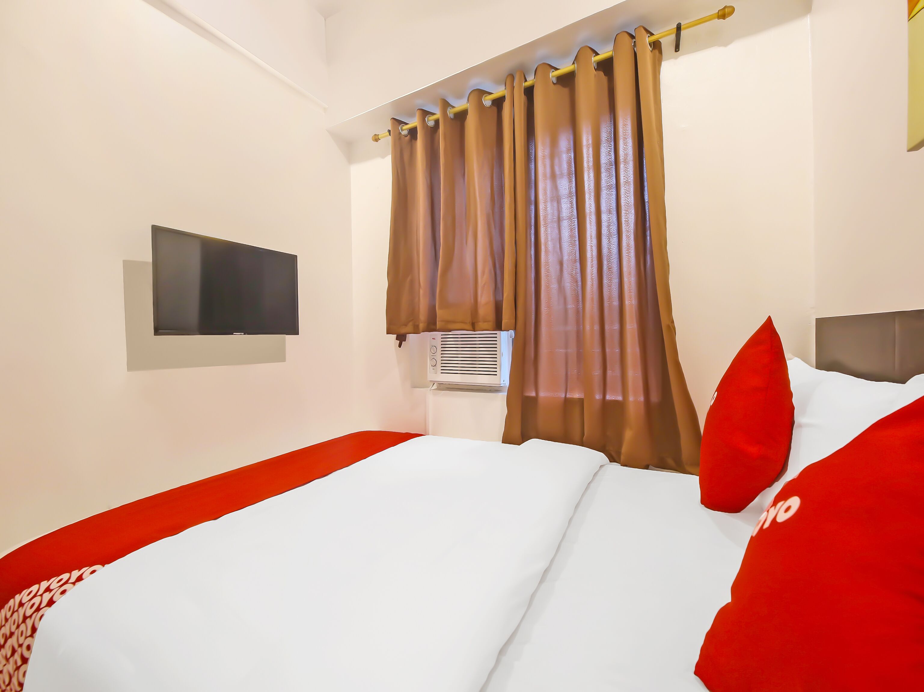 A hotel room with a bed, a flat-screen TV mounted on the wall, an air conditioning unit, and brown curtains.