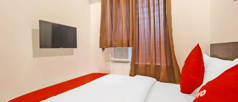 A hotel room with a bed, a flat-screen TV mounted on the wall, an air conditioning unit, and brown curtains.