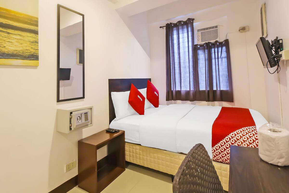 Ein Hotelzimmer mit einem Bett, einem Nachttisch, einem Spiegel, einem Fenster mit Vorhängen und einem an der Wand montierten Telefon.