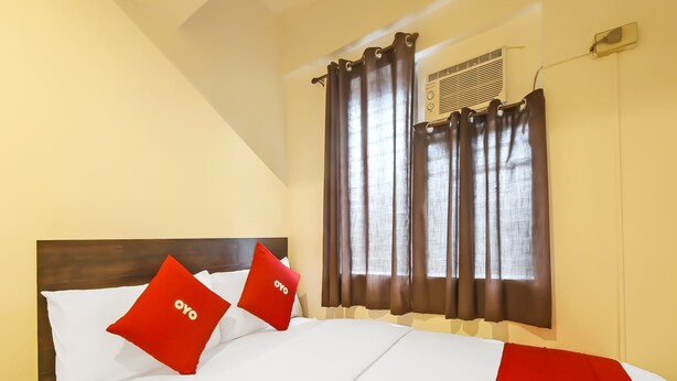 Habitación de hotel con cama, dos almohadas rojas con el texto "ovo", una manta roja, un cabecero de madera, una ventana con cortinas y un equipo de aire acondicionado.