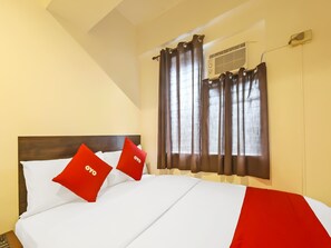 Une chambre d’hôtel avec un lit, deux oreillers rouges ornés du mot « ovo », un plaid rouge, une tête de lit en bois, une fenêtre avec des rideaux et un appareil de climatisation.