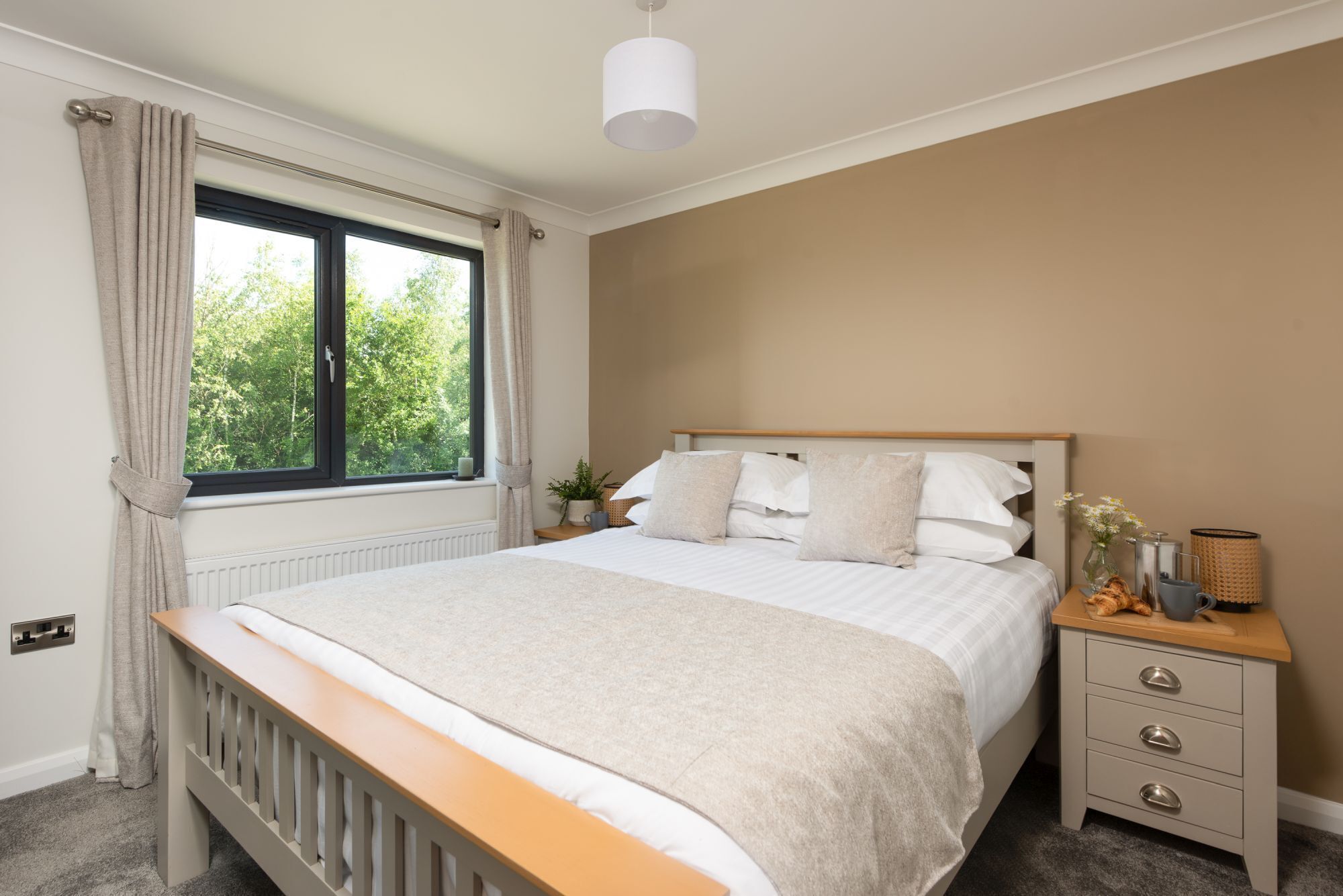 A bedroom with a large bed, a bedside table, a nightstand, a window with curtains, and a view of greenery outside.