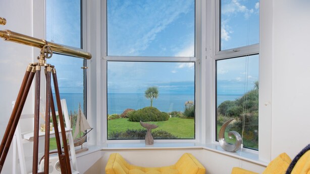 A room with a large window overlooking a coastal landscape, a telescope, and a yellow armchair.