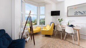A bright living room with a large window, a tripod, yellow chairs, a dining table, and a TV on the wall.