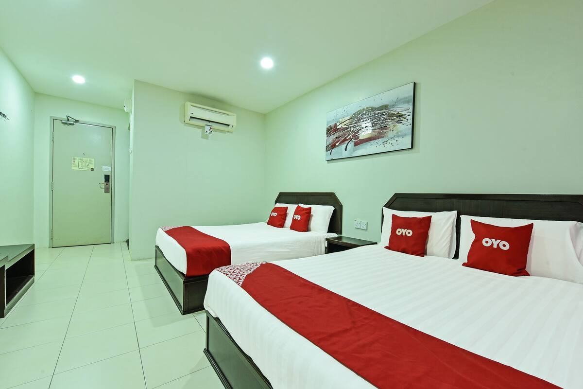 Habitación de hotel con dos camas, cada una con cubrecama y almohadas rojas y blancas. Se observa un cuadro en la pared, una puerta con un cartel y un equipo de aire acondicionado.