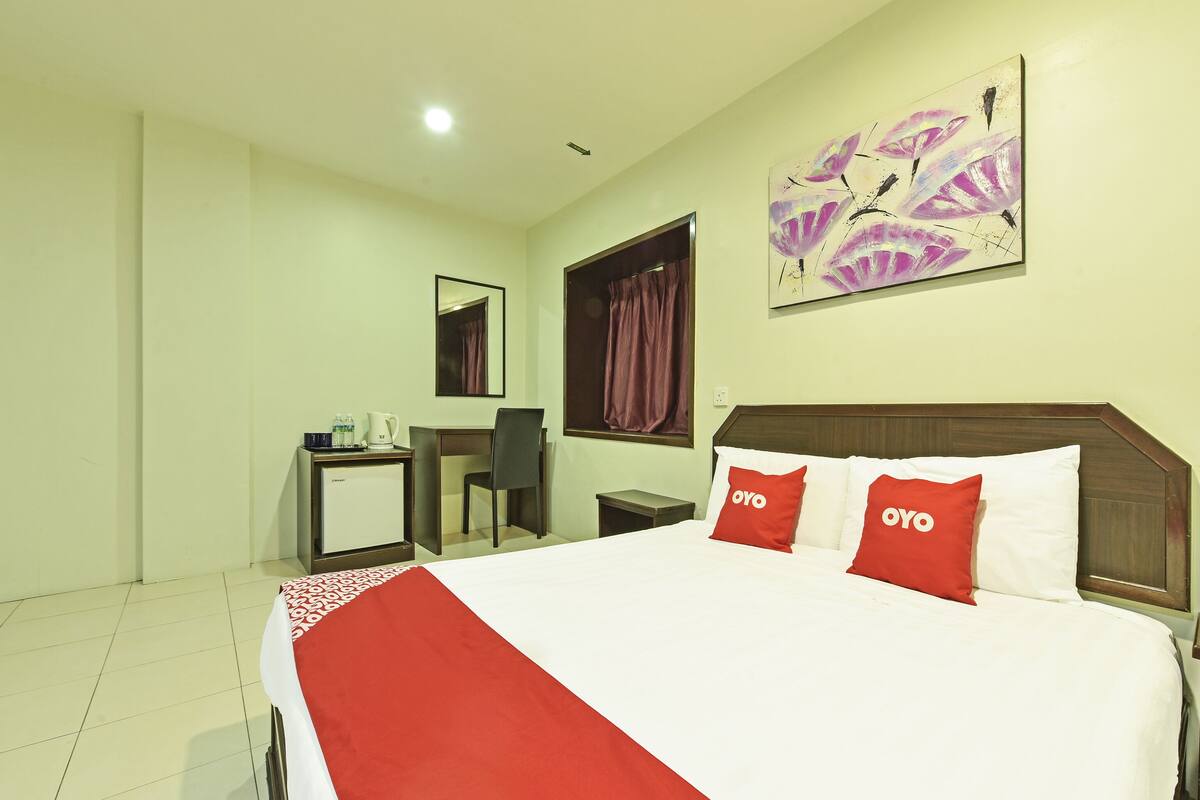 Habitación de hotel con cama, escritorio, silla, pequeña mesa, cafetera y un cuadro en la pared.
