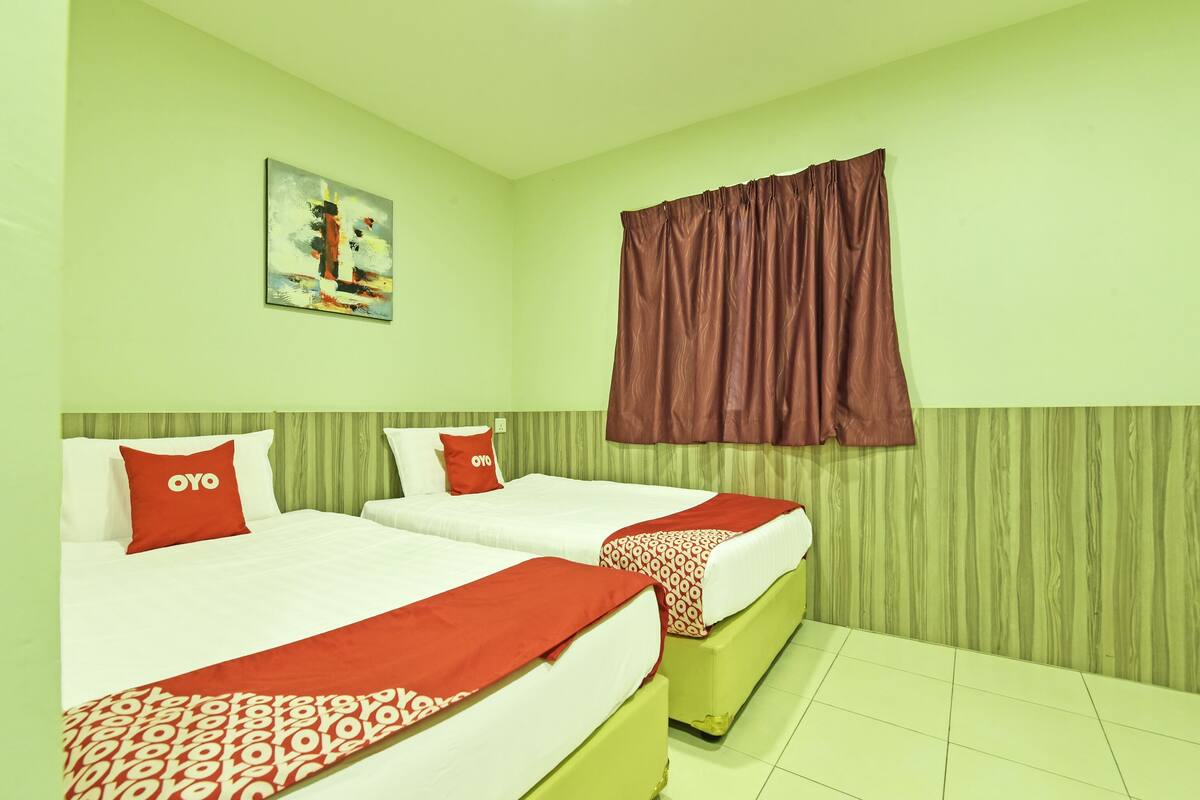 Habitación de hotel con dos camas, paredes verdes, un cuadro en la pared y una ventana con cortinas marrones.