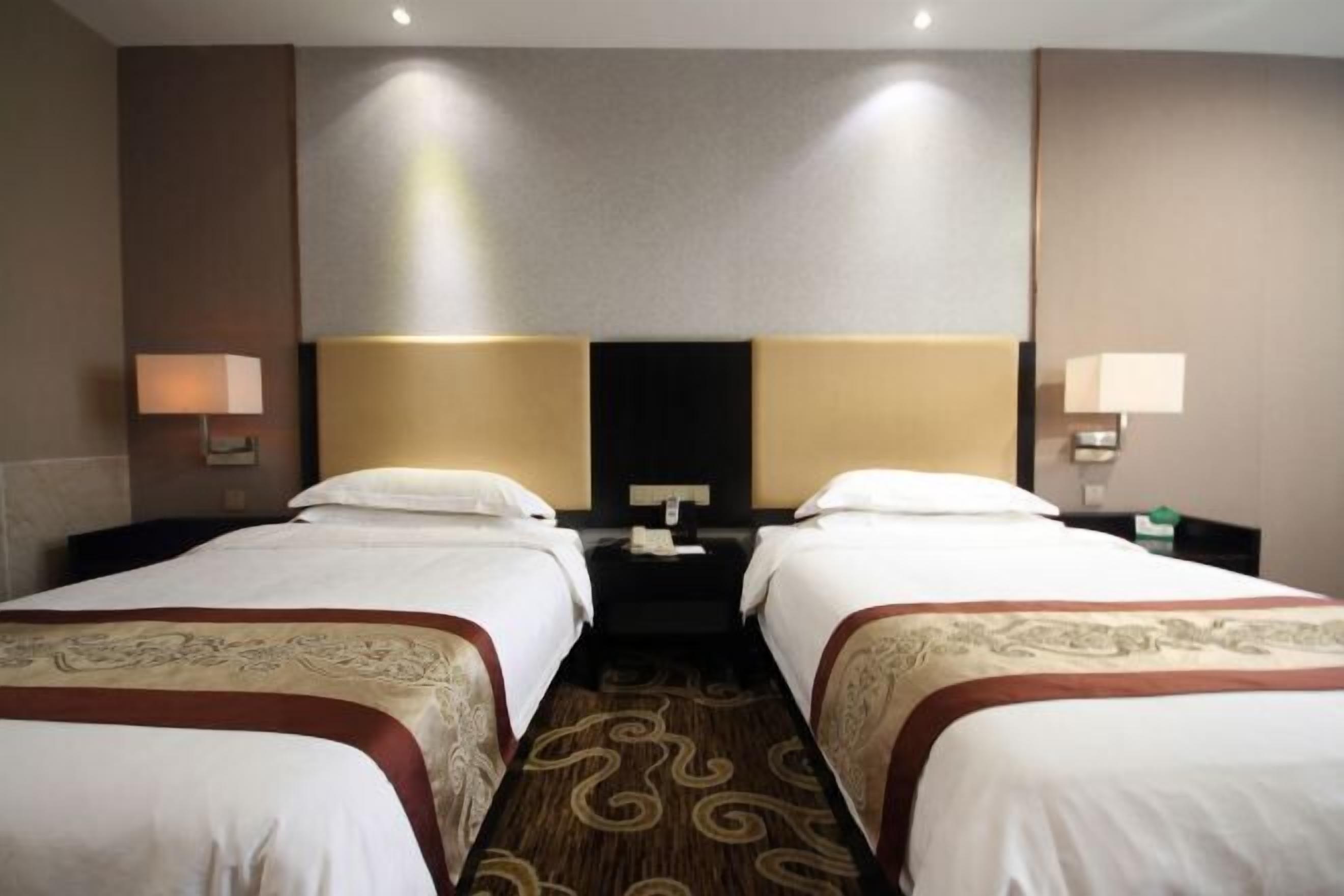 Habitación de hotel con dos camas, cabecera oscura, mesita de noche con lámpara y piso alfombrado.