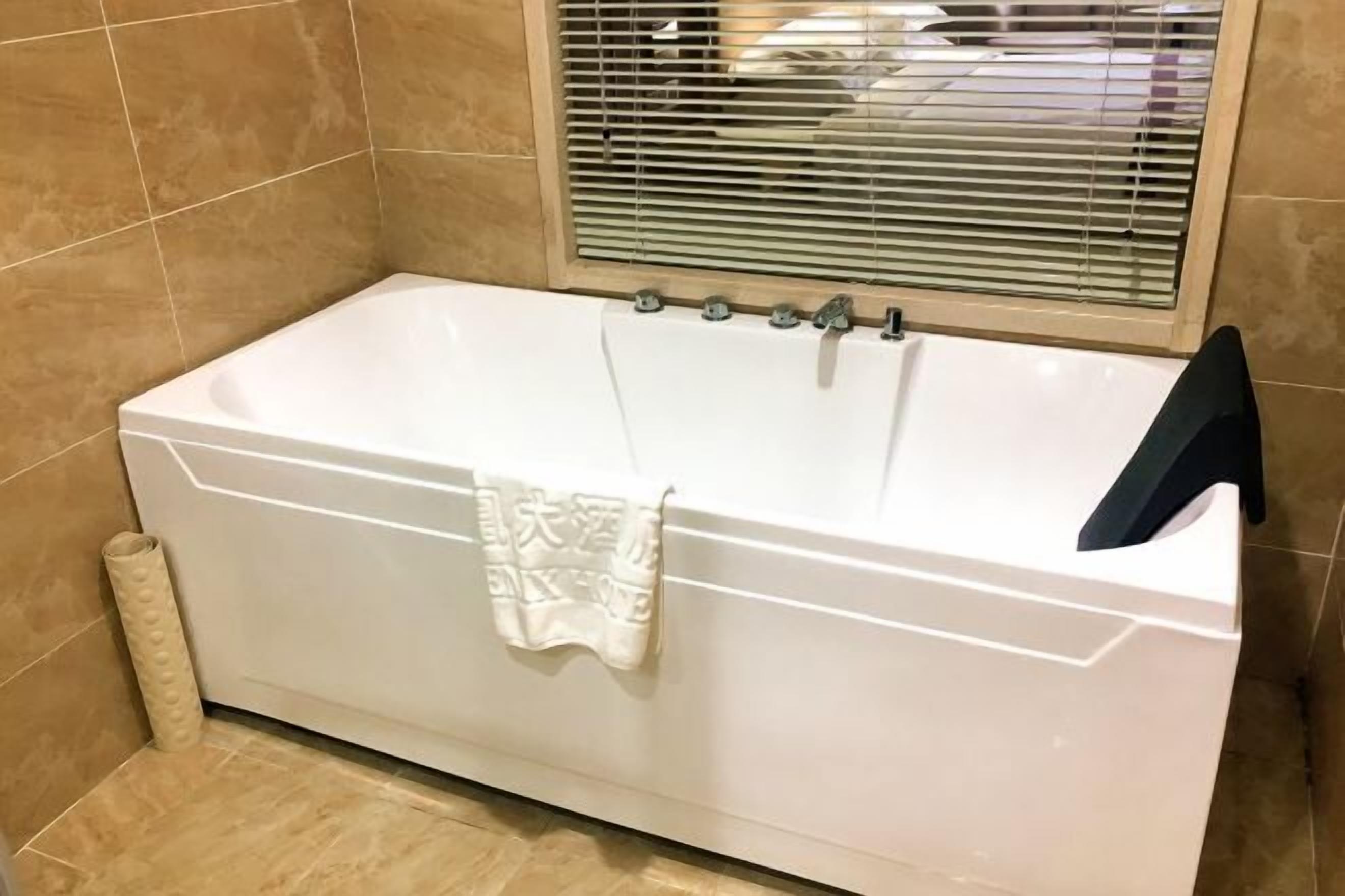 A white bathtub with a towel hanging on it, a window with blinds, and a roll of toilet paper on the floor.