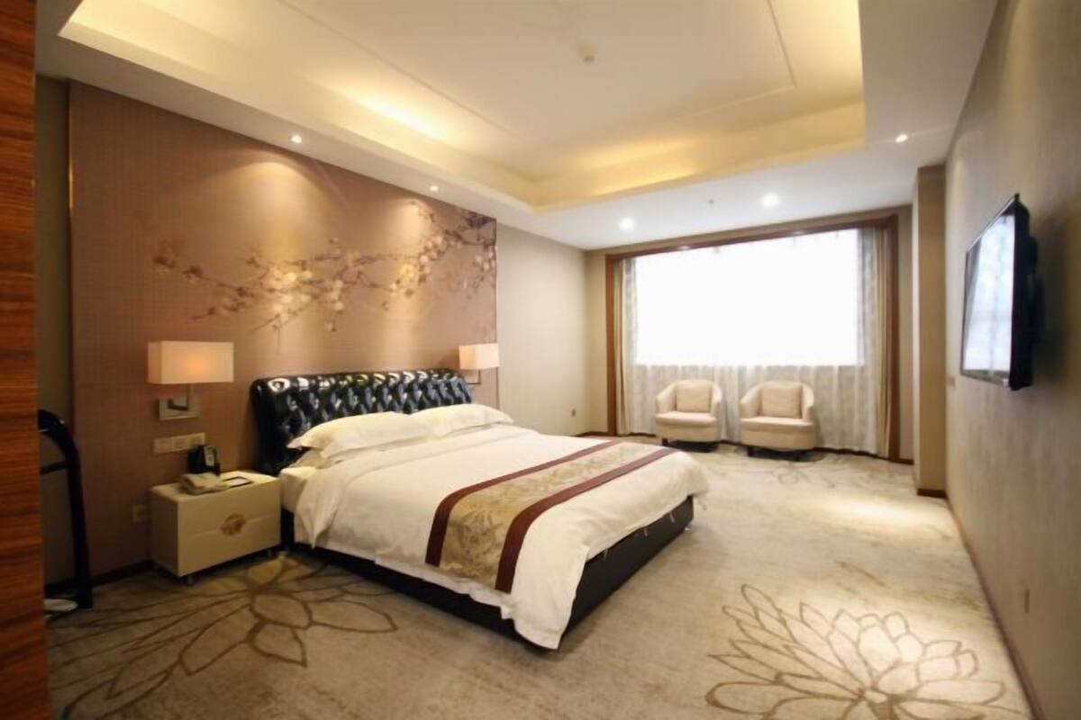 A hotel room with a large bed, a desk, two chairs, a TV, and a window with curtains.