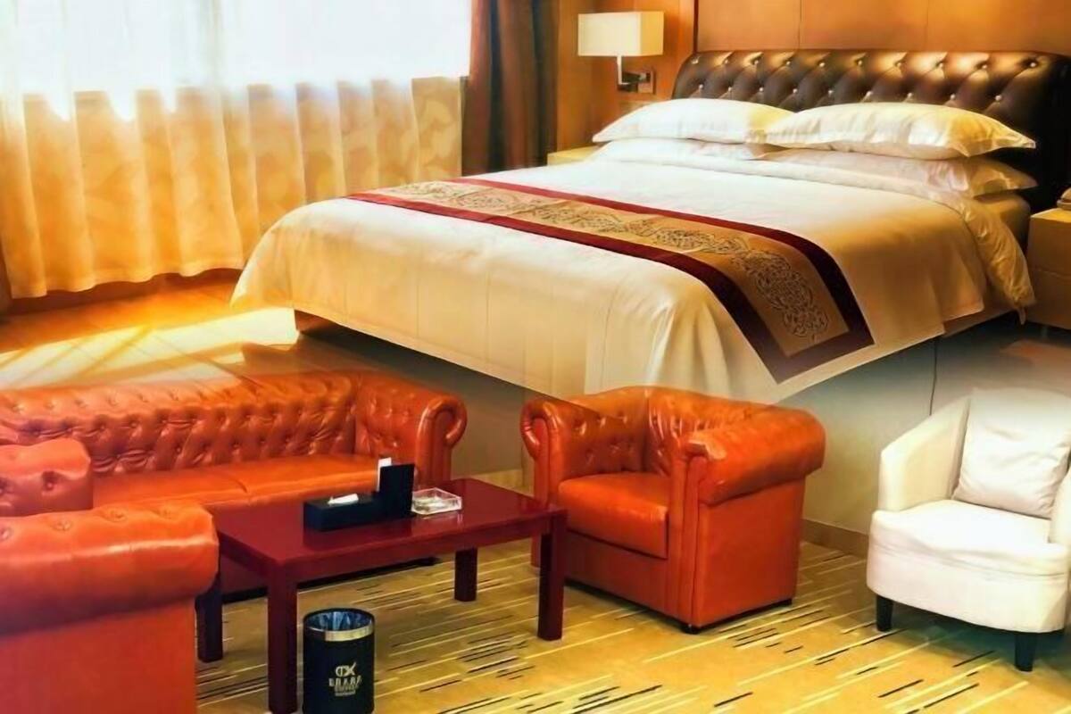 A hotel room with a bed, two leather sofas, a white armchair, a wooden coffee table, and a lamp.