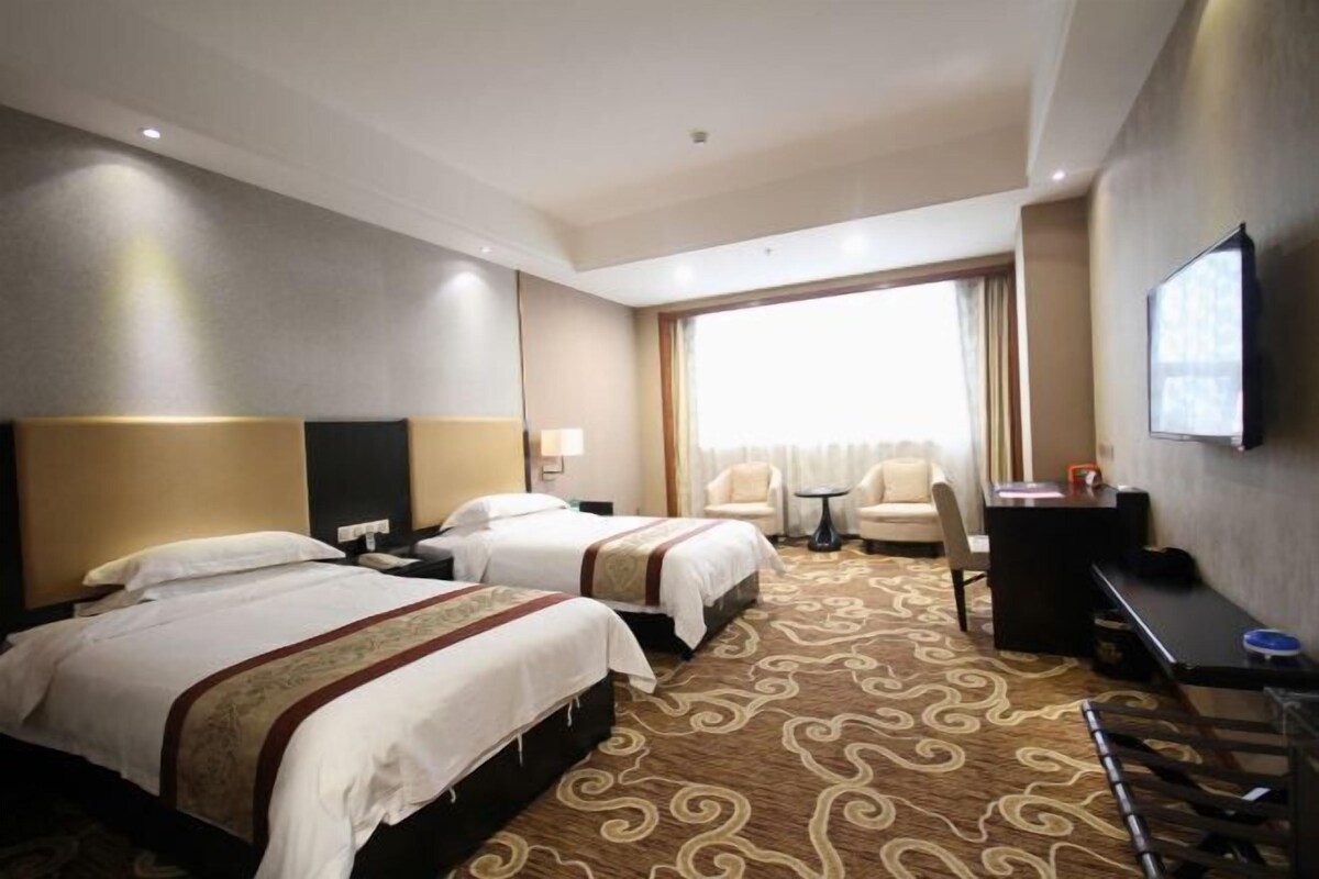 A hotel room with two beds, a desk, a chair, a TV, and a large window.